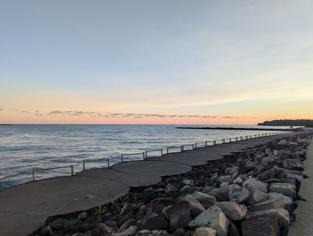 A light pink and blue sky with wispy clouds over a lake as the sun rises. In the foreground are rocks and a walking path with rails.