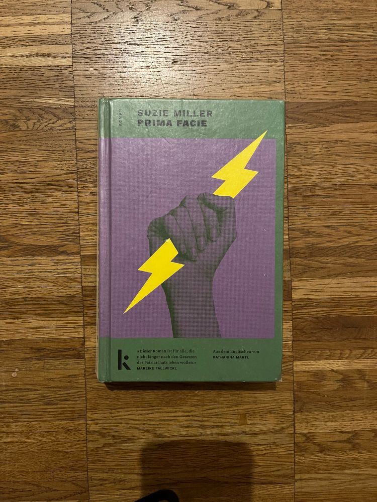 Holzboden, auf dem das Buch „Prima Facie“ von Suzie Miller liegt. Der Rand des Covers ist grün, mittig eine große lila Fläche mit einer Hand (einer weiblich gelesenen Person), die einen Blitz in ihre Faust geschlossen hat