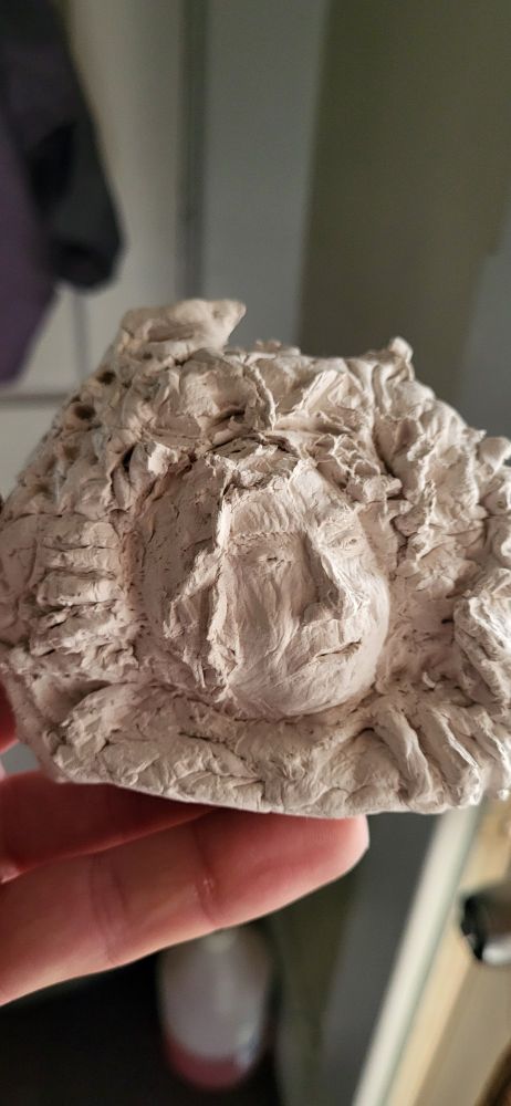 A small 3x3 inch sculpture of a face emerging from a bush/thicket with a hand on either side of their face, as if pulling themself out (expression on face is on of concern/struggle)