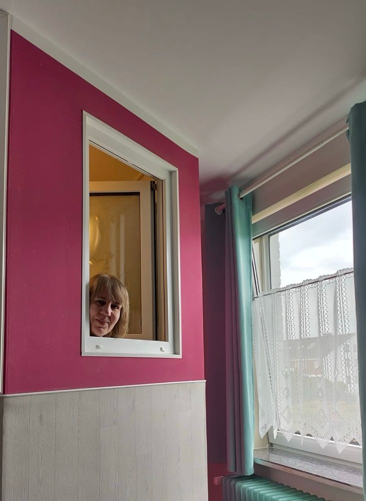 Foto von mir, wie ich aus einem kleinen Fenster gucke, das in ein Hotelzimmer reinführt 