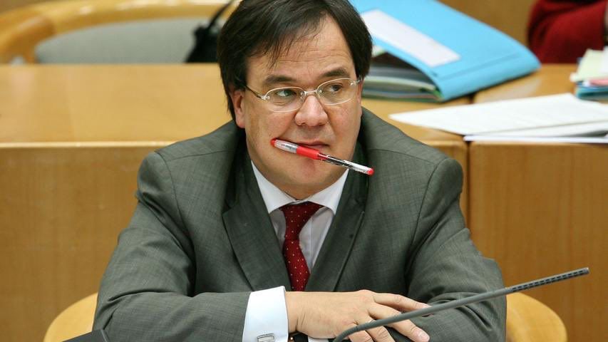 Ein jüngerer Armin Laschet hat sich einen Kugelschreiber an die Lippe geklemmt oder hält ihn mit den Zähnen. Er trägt Anzug und schaut gelangweilt drein.