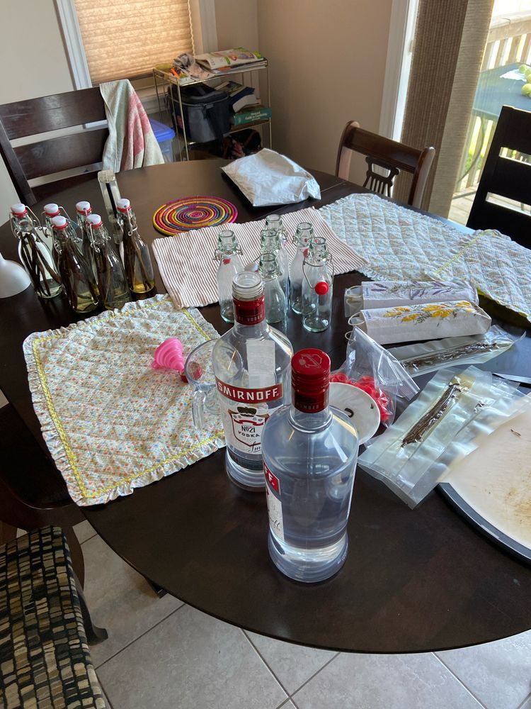 A kitchen table with small bottles of vodka and vanilla beans on them. Beside them sit the same bottles, only empty. Two large bottles of Smirnoff vodka sit in the foreground.