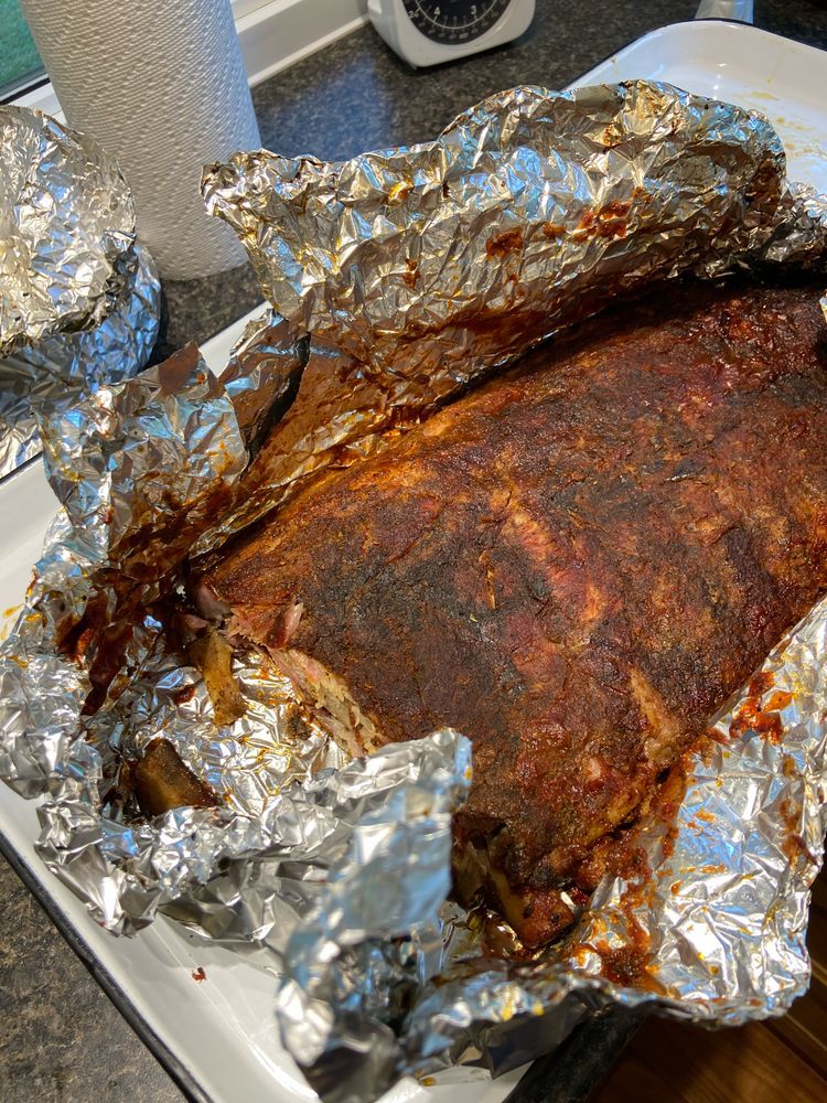 A cooked pork side rib in a tin foil wrap, coloured darkly from cooking. 
