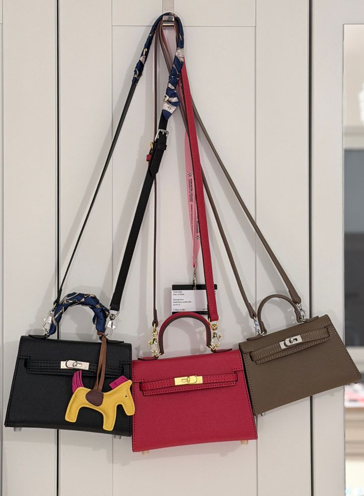 Three handbags hanging on a white door with panels. All with shoulder straps. 
Left: Black Kelly Mini type bag, black thread, palladium colored steel hardware.
Handle silk wrapped, blue colored print ornaments. 
A yellow horse of leather with pink mane and tail.
Middle: red Kelly Mini type bag, red thread, golden colored brass hardware.
Right: brown Kelly Micro type. Palladium colored steel hardware. 
