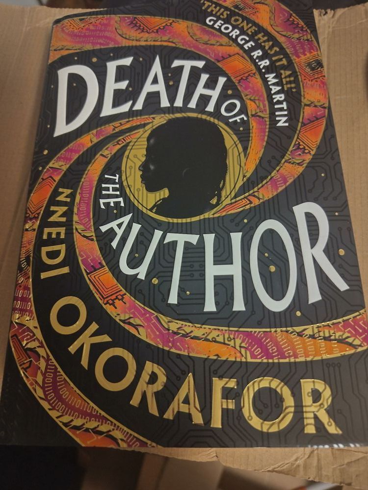 Cover of Death of the Author by Nnedi Okorafor. Features a stylized silhouette against a swirling, patterned background. Blurb by George R.R. Martin , 'This one has it all!', is visible at the top. Book rests on a cardboard surface.