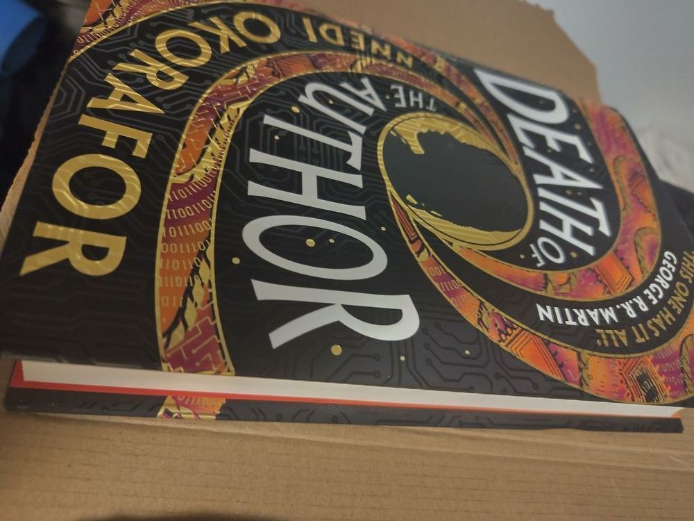 A book titled The Death of the Author by Nnedi Okorafor. UK edition (not sprayed pages)  is pictured laying in an open cardboard box. The cover design features intricate patterns and bold, swirling typography in black, gold, and red tones.