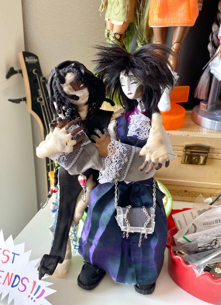Photo of my Stop Motion puppets DU and Josephina, standing next to each other in a friendly embrace. DU stands on one leg as he leans onto Seph with one arm around her shoulder while Seph reaches out for his other hand standing straight with the other arm crossed over her chest. They stand on a white shelf