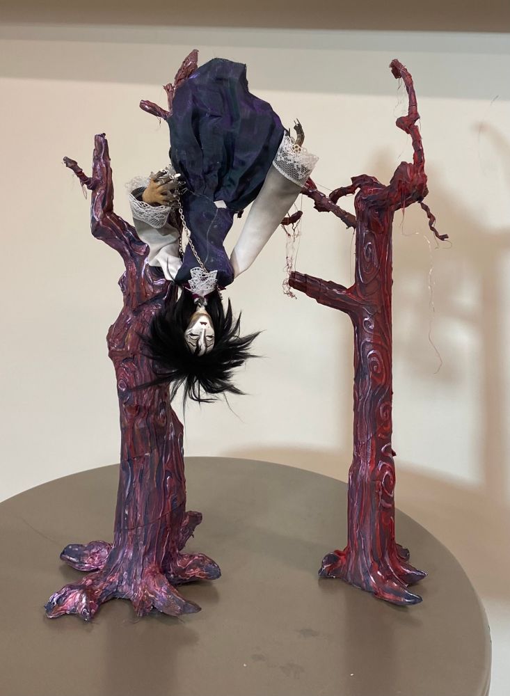 Babs’ puppet Seph, hangs with her legs over the branch of a dead tree. The trees are made from cardboard, hot glue, and paint for texture. Ones is thicker and juts out, while the other is thinner with more swirls in the bark. The thicker one is purple tinted while the thin is reddish, both are barren and dead