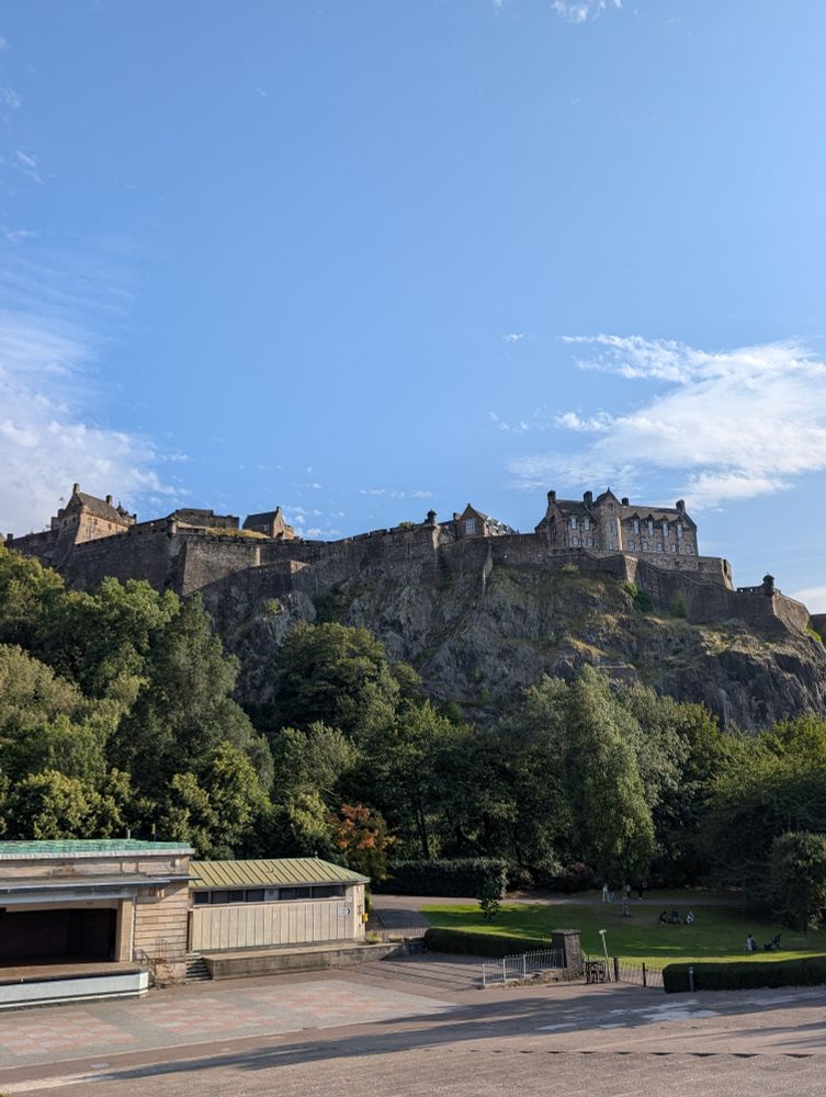 View of Edinburgh Castle from a park below. The castle is perched upon the top of a rocky hill, and the base of the hill is surrounded by trees.
