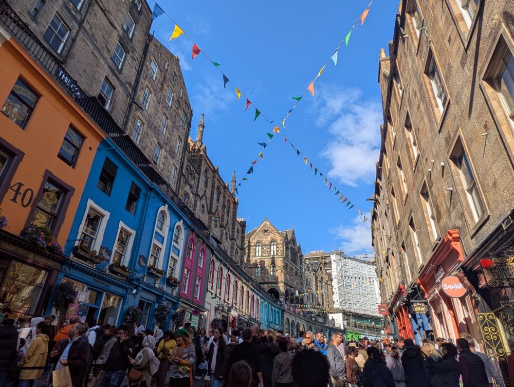 A crowded pedestrian street in central Edinburgh, with decorative banners strung over the street. The buildings are painted in various colors.