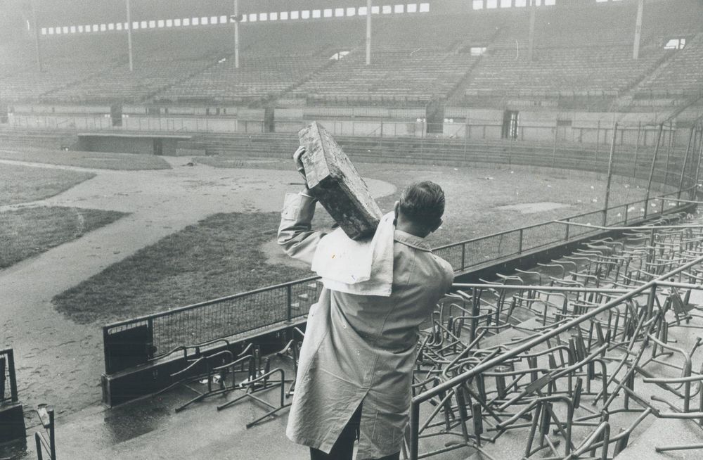 A photo of Maple Leaf Stadium in its final days. It's empty and looks abandoned. Seats have been ripped out. The field is in disrepair. It's raining and wet. The man who bought homeplate for $5 as a souvenir — Len Andrews — is walking away with it, holding it up on his shoulder, a towel under it to protect his coat.

(Image: Toronto Public Library)