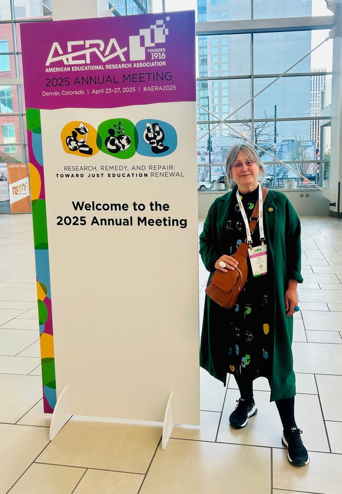 A grey haired woman in green coat and smiling stood next to the AERA conference welcome sign.