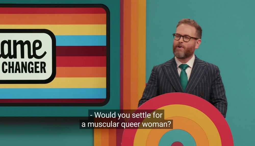 Host Sam Reich of the Dropout show, Game Changer. He is wearing one of his signature pinstripes suits, standing behind a colourful podium on the Game Changer set. The closed captioning reads

“Would you settle for a muscular queer woman?”