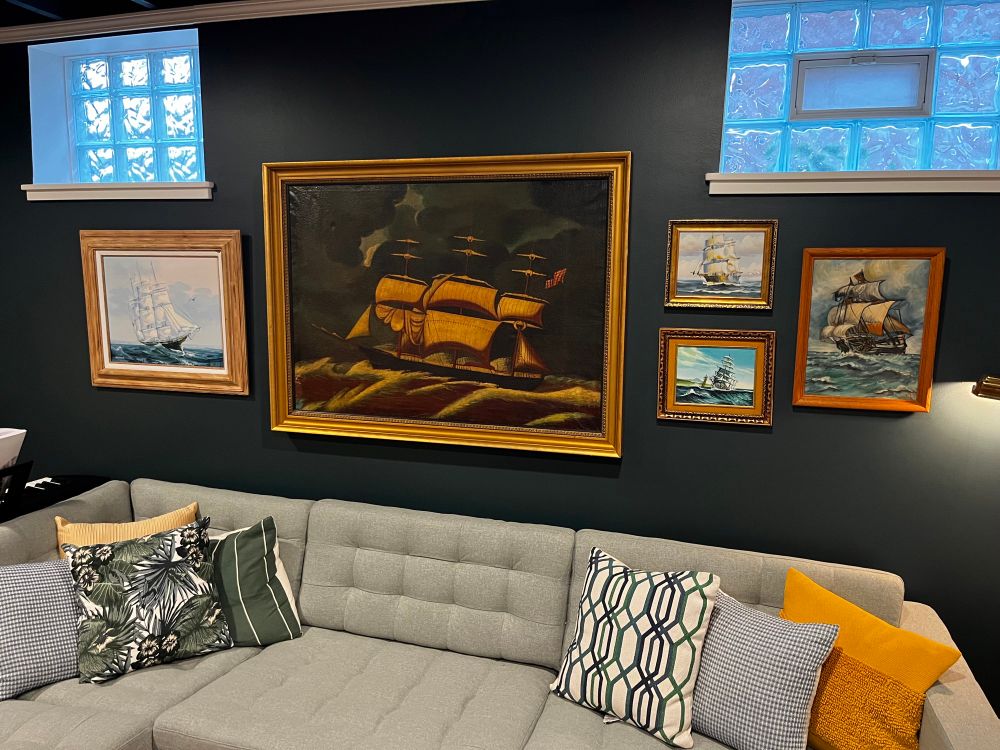 A gallery wall in a basement living room. 5 paintings of old ships of various sizes in gilt frames hung on a deep teal wall above a sage green room couch.