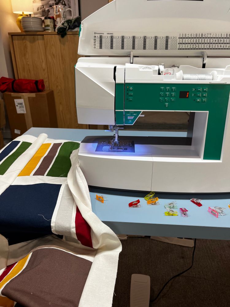 A progress picture of a quilt topper being assembled at my sewing machine with solid green, brown, yellow, red, blue, and grey blocks separated by an off-white fabric