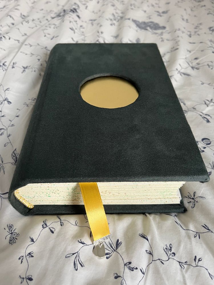 a thick, hardcover book bound in dark green velvet with a shining, metallic, golden circle cut out  of the centre. the cream edges of the paper are speckled with green and gold, and there is a gold ribbon bookmark hanging out of the bottom of the book