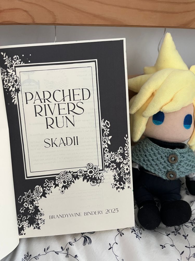 the book open to its title pages, which reads PARCHED RIVERS RUN by skadii and BRANDYWINE BINDERY 2025 below. the text is black inside of a cream-coloured box, and the box is on a pure black background, surrounded by flowers. next to the book is small plush version of cloud strife, with pointy yellow hair and round blue embroidered eyes