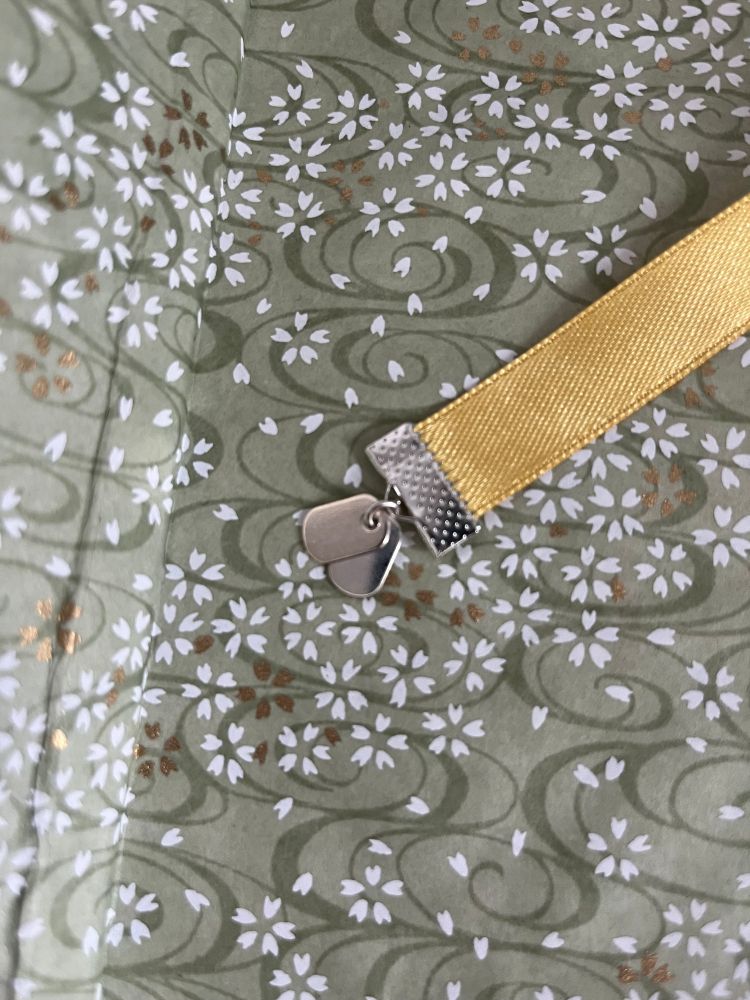 tiny silver dog tag charms on the end of a gold ribbon, against a pale green background with darker green swirls and white and gold flowers