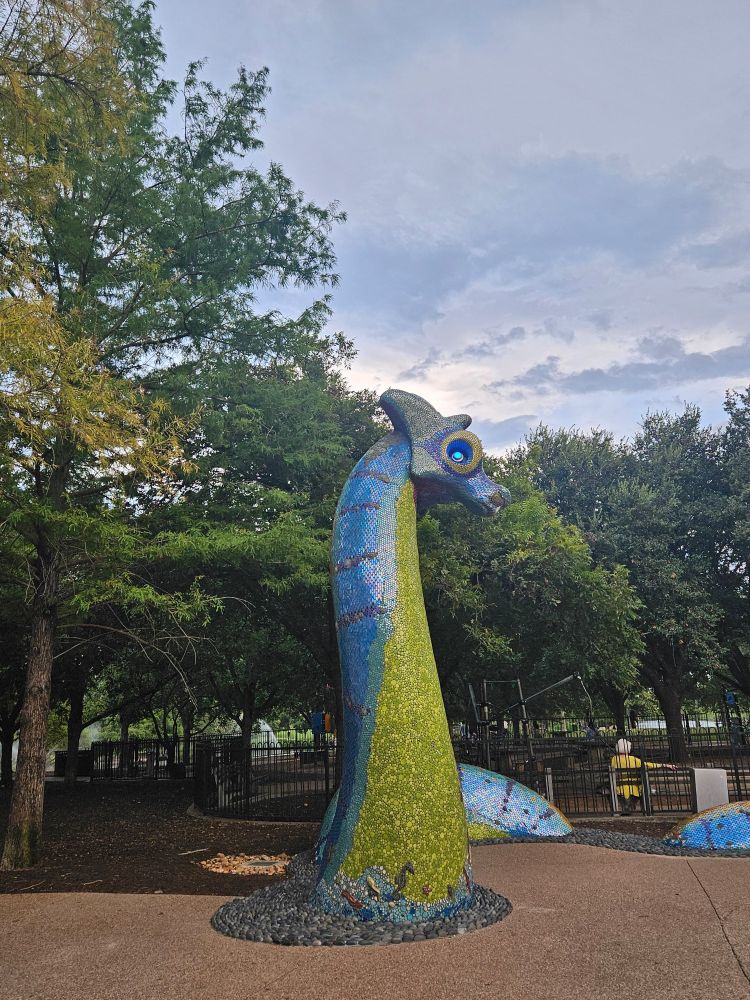 A sea monster statue with a glowing blue eye, captured in profile at the edge of Mueller Lake Park. The surrounding trees match its chartreuse belly, while its blue back is mirrored by the cloud cover overhead. Just past the statue, a woman in yellow sits on a bench in a playground, her back to the camera.
