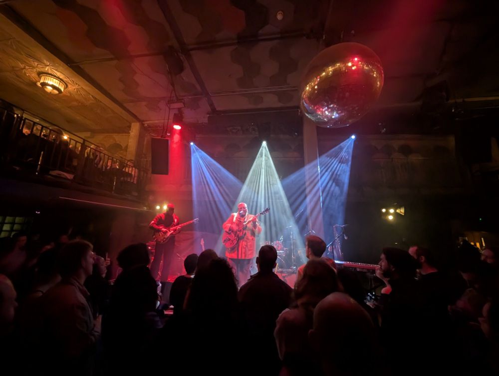 Two people with guitars on stage with one person singing into a microphone. Lights illuminating the singer. Wide angle