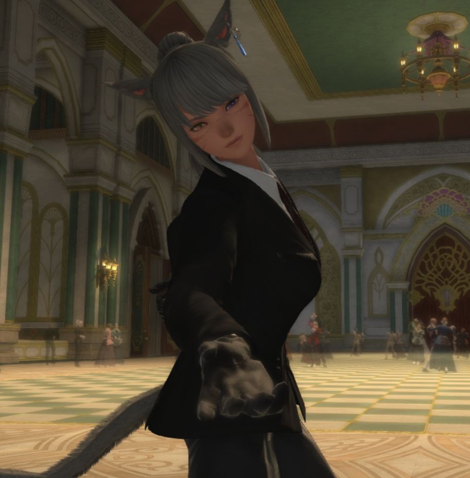 Kara extends her hand towards the viewer. She's standing in a ballroom, wearing a suit.