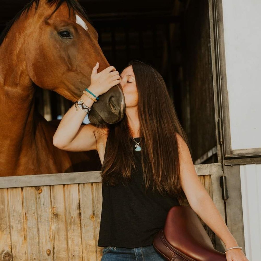 Dark brown long haired female kissing a bay warmblood horse while holding a saddle 