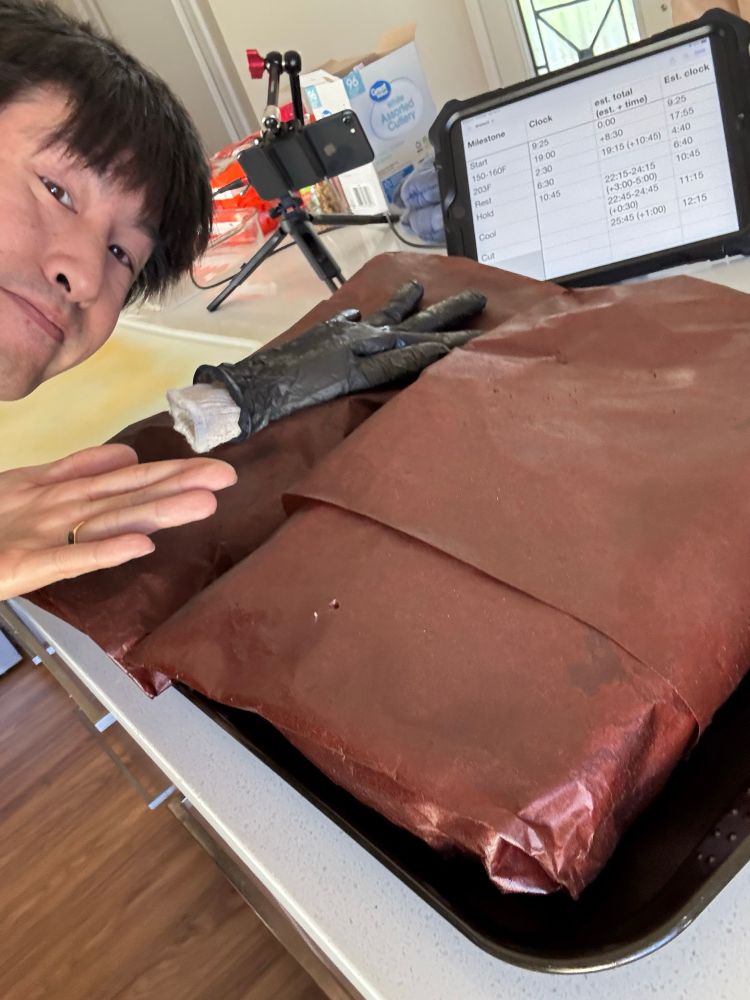 Wrapped briskets sitting on a counter with a filled spreadsheet of cooking milestones displayed behind it