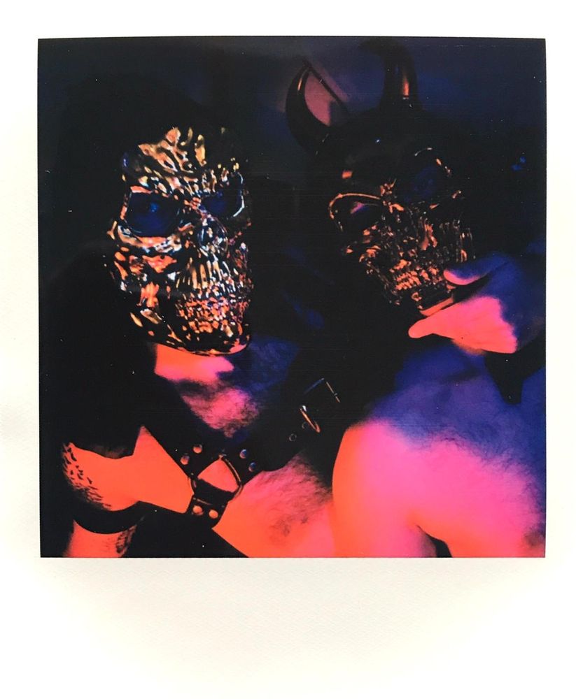 Krosscumback and Laddie Macbeth lie together in repose wearing gold skull masks. Their torsos are lit in soft pinks, oranges, and purples. 