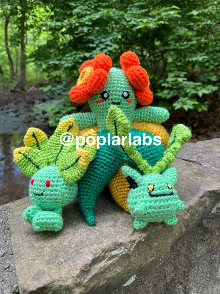 Three crocheted amigurumi based on different Pokémon characters sit on a stone bridge overlooking a creek. The forest in the background is lush and green, like these plushes. The largest one is Bellossom. The smaller ones are Shiny Oddish and Shiny Hoppip.