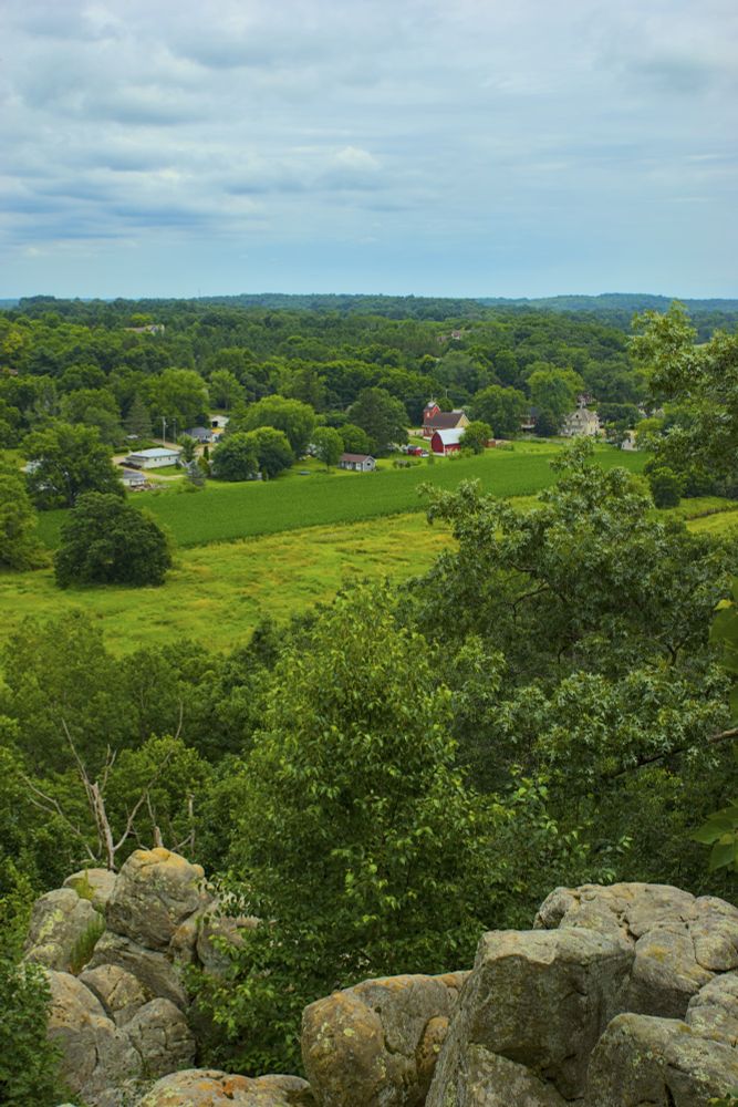 image of tree-lined landscape with red barn and field in middle and rocks in foreground