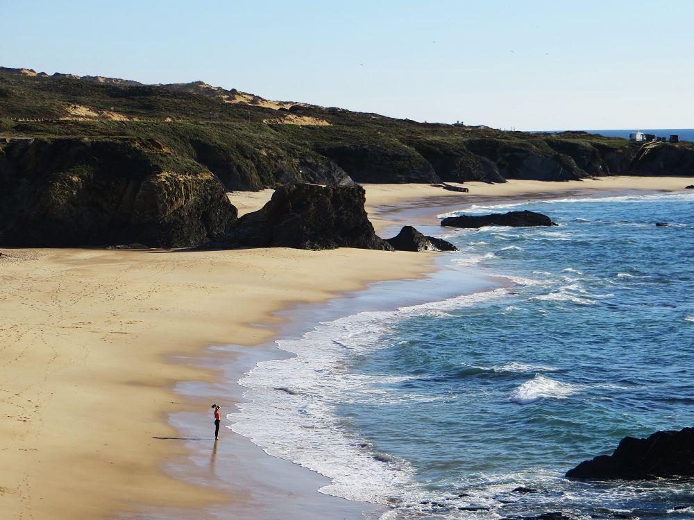 Woman stands alone near the surf on Furnas Beach, SW Portugal: a large beach separated by rocks at high tide. By Sonja Holverson @SonjaSwissLife
