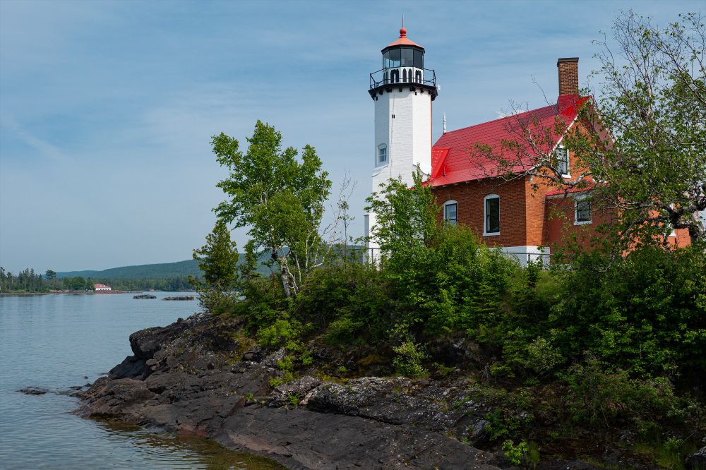 A white painted lighthouse stands on top of a rocky shore. The attached keepers house is in unpainted brick, and there are red roofs on both. In the distance the old Life-Saving Station boat house can be seen. Taken on a glorious summers day under blue skies.