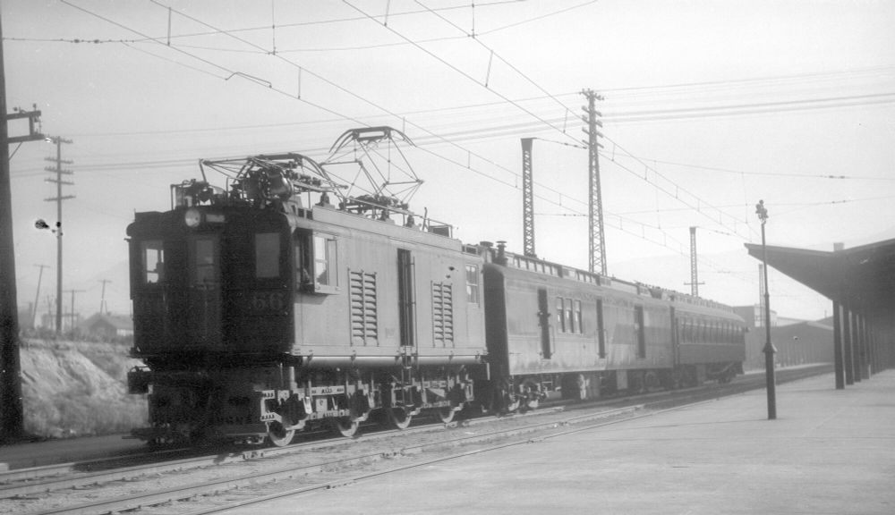 A black and white photo of a boxy B-B boxcab electric locomotive pulling a 2 car train of a heavyweight railway post office/baggage car and a coach. A station platform is visible at the right.