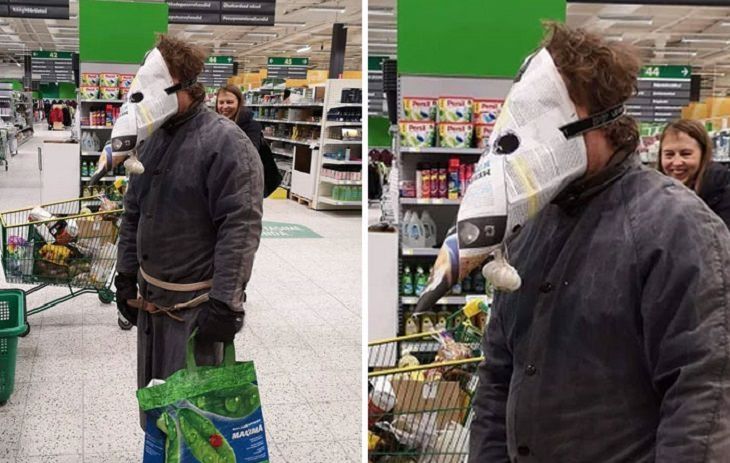 A person is standing in a grocery store. Behind them is an aisle end cap as well as a person with a shopping cart, suggesting they are standing in a spaced out checkout line. Over their face is newspaper rolled into a cone shape, two eye holes cut out and a full head of garlic dangling from one side.