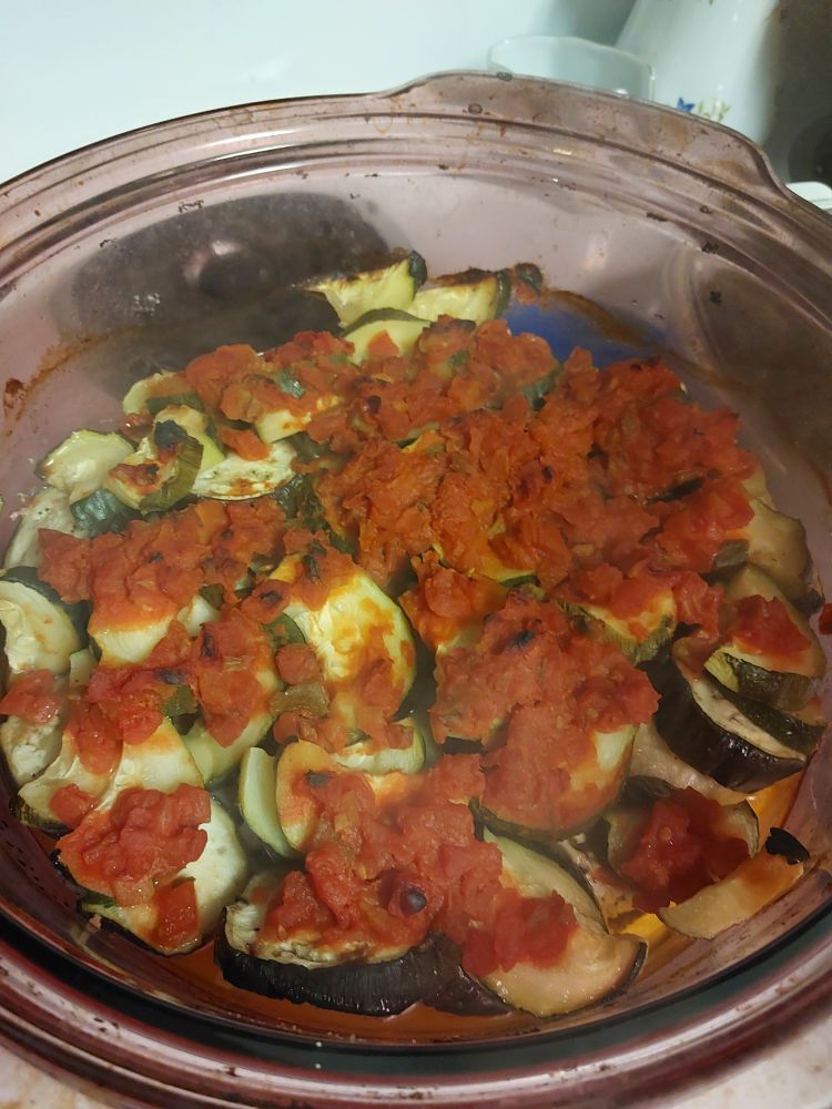 A purple glass pot with baked vegetables in it. On top are clumps of diced tomatoes, followed by zucchini and eggplant slices. Hidden by the above are chicken leg quarters, layered into the pot whole. Not pictured: wilting but still good tarragon in the fridge, also cheese that will go on the vegetables later.