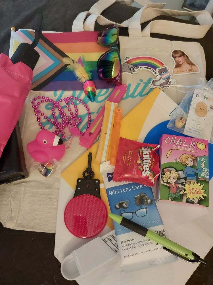 Pride swag! Laying on top of an off-white canvas tote: pride flag, sunglasses, two pride cat stickers, a Taylor Swift sticker, proud potato in a sheer white drawstring bag, white yellow and blue mini tote, umbrella in a pink sleeve, black sharps box, flamingo pink bead necklace and rubber squeaky toy, fan in pink casing, heart shaped pride pin, orange drink packet and bubble stuff tube, chalk, pouch of skittles, red reflective light up keychain, mini lens care kit, green and black click pen, slide measuring spoon, and a rainbow mouse cat toy with feather tail. Not pictured: cane freshly wrapped in iridescent tape that didn't make it to Sunday's events because lemon flavored connective tissue happened and my wrist was doing worse than the foot. The gremlin loves her new mouse.