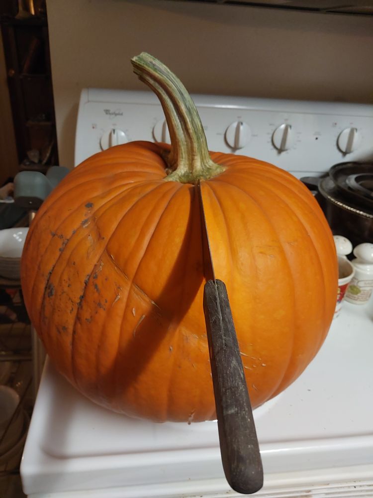 A cleaver is stuck into a large jack o lantern pumpkin, handle pointed toward the camera. It is sitting on a white stove. Behind to the right are salt and pepper shakers, a mug, and a pan and lid. Leaning against the stove to the left is a crutch.