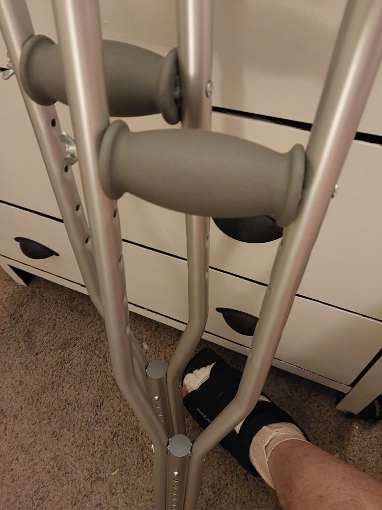 Crutches leaning on a cream colored dresser with dull bronze toned shell handles. Dangling above the brown speckled floor between the crutches and dresser is a wrapped foot. Gauze pokes out from under ace bandage and covers all but the big toe. Over the ace bandaging is a short black fracture shoe.