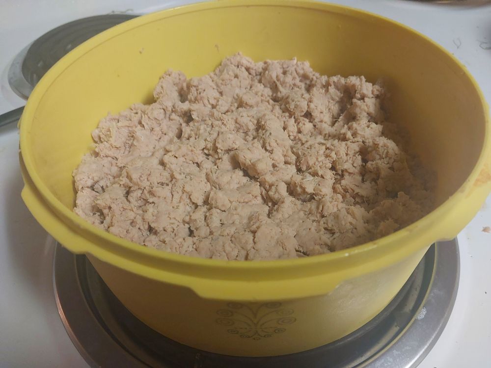 A round piece of yellow vintage Tupperware is sitting on a burner coil on a white stovetop. In the Tupperware is unseasoned cooked chicken paste.