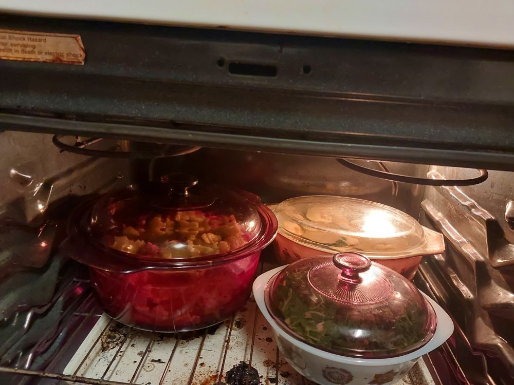 Two baking dishes and a glass stock pot in the oven. On the left is the stock pot with beef, swiss chard stalks, banana pepper, and turnips. Rear right is an Early American brown oval pyrex baking dish with large chunks of acorn squash, the skin still on. Front right is a vintage round glasbake dish with chopped swiss chard leaves with salad turnip greens buried underneath, topped by a mismatched purple pyrex lid. Below them is an off-white cookie sheet permanently on spillover catch duty.