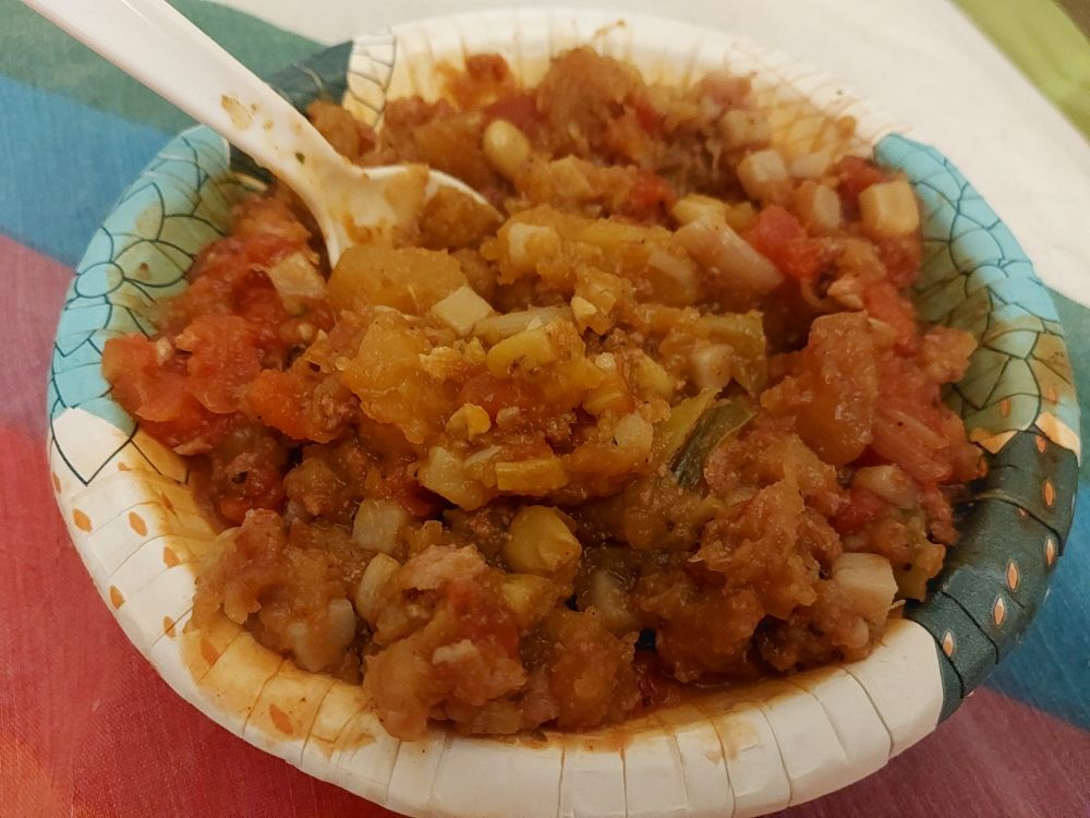 A blue and white printed paper bowl sits on a rainbow striped sheet. In the bowl is roughly mashed pumpkin with corn, potato, tomato, leek, and canned meat.