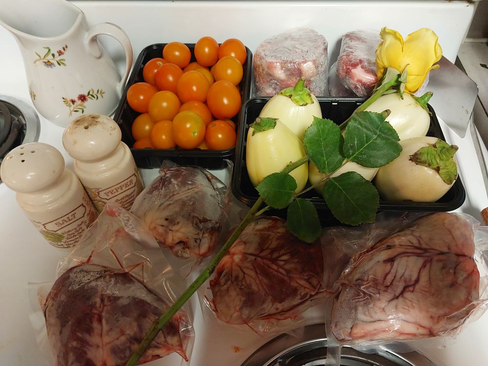 Meat and vegetables sitting on a white stove. From back to front: a black tray of orange mini tomatoes, lamb neck slices, a black tray of garden eggs, and four lamb hearts. A yellow rose is laid diagonal across the top. To the left is a white with floral print cream pitch and a set of vintage Avon salt and pepper shakers, and to the right is a metal spatula with wooden handle.