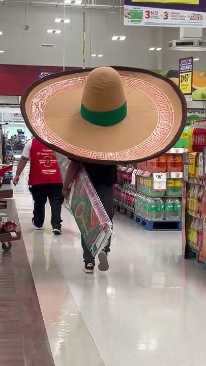 A nearly aisle-wide sombrero, going down the main pathway of a grocery/department store. Under it is a figure carrying plastic wrapped colorful paper. The sombrero is following a store employee wearing a red vest and passing displays of soda to its right.