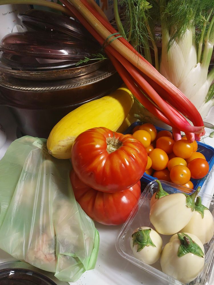 A stack of glass and metal stock pots sit on the rear burner of a stove, a mound of vegetables in front of them. From back to front: fennel, rhubarb, two yellow squashes, orange mini tomatoes, two beefsteak tomatoes, lamb hearts in a translucent green bag, garden eggs.