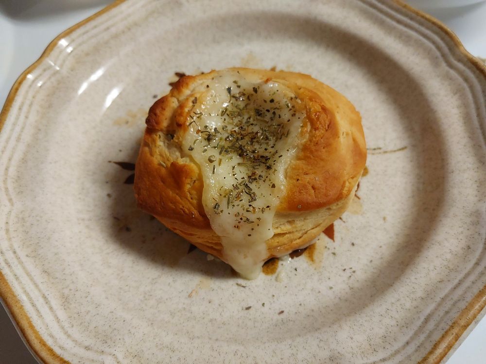 A golden brown biscuit is sitting on an old plate, fontina cheese melted and spilling out from a hollow in the center with green herb seasoning sprinkled on top. The plate is gray speckle on cream with scalloped edges and tan along the rim.