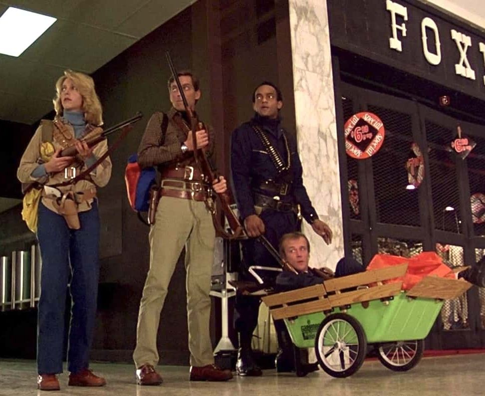 From the movie Dawn of the Dead (1978). Two men and a woman are standing in a mall, watching for zombies. There is a fourth person with them, sitting in a lime green rolling cart due to his injury from getting bit.