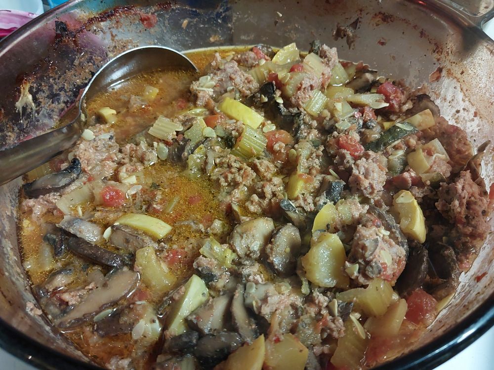 A clear brown glass stock pot with soup in it and bits stuck to the sides from baking in the oven, with a silver colored ladle stuck in and tipped to the left. Breakfast sausage, zucchini, yellow squash, tomatoes, mushrooms, and celery make up most of the topography with brown broth and golden liquid fat shimmering at the surface in a few places.