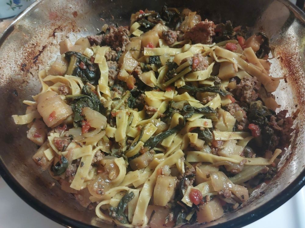 Soup fixins baked in a brown glass stock pot, sitting on a white stovetop and yearning for broth. Behind is a purple glass pot.
In the pot are beef, homestyle egg noodles, radish greens, swiss chard leaves, kohlrabi, tomato, and onion.

