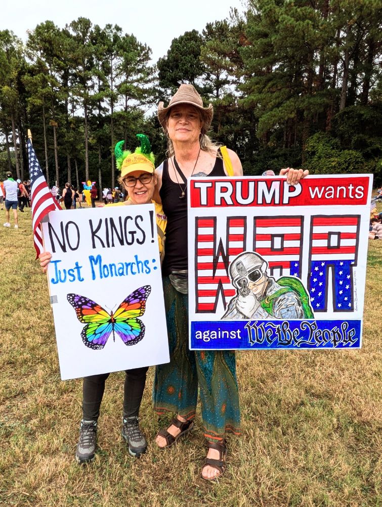 Two protesters hold up signs at the no. Kings protest in Atlanta on October 18th to 2025. One sign reads no Kings just monarchs and shows a picture of a butterfly. The second sign says Trump wants war against me the people.