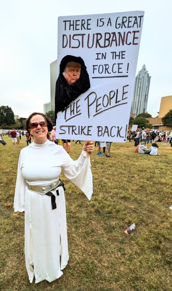 A woman press dressed as princess. Leah holds up a protest sign that says there's a great disturbance in the force. The people strike back at the no. Kings Protestant land on October 18th 2025